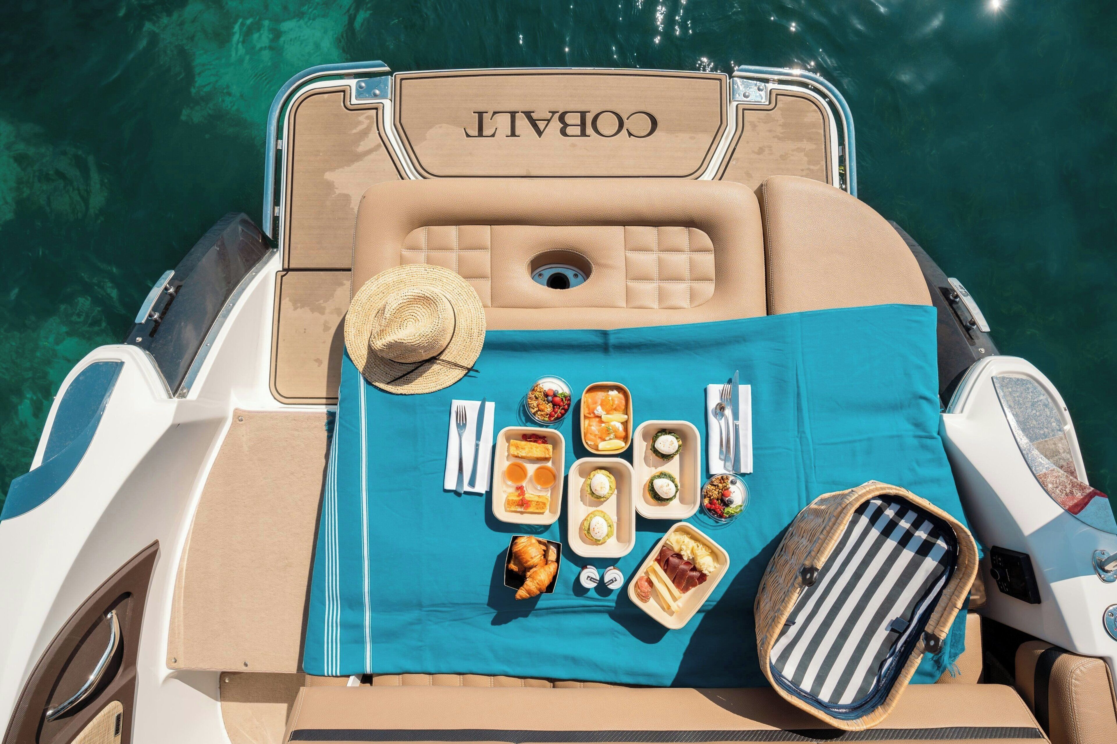 Un bateau dont l’intérieur est bleu et beige, un chapeau et une table dressée avec de la nourriture et des boissons.