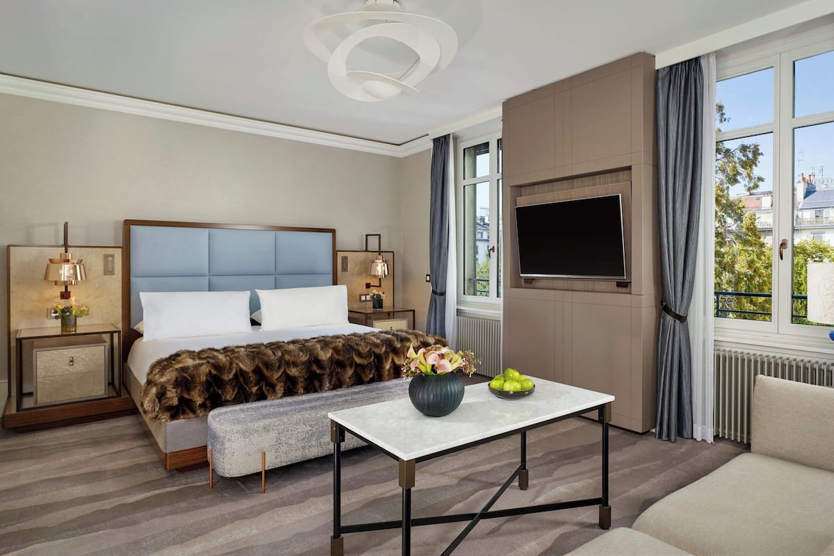 A hotel room with a large bed, a flat-screen TV, a small table with a vase, and a view of the city through the window.