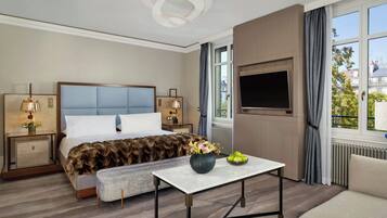 A hotel room with a large bed, a flat-screen TV, a small table with a vase, and a view of the city through the window.