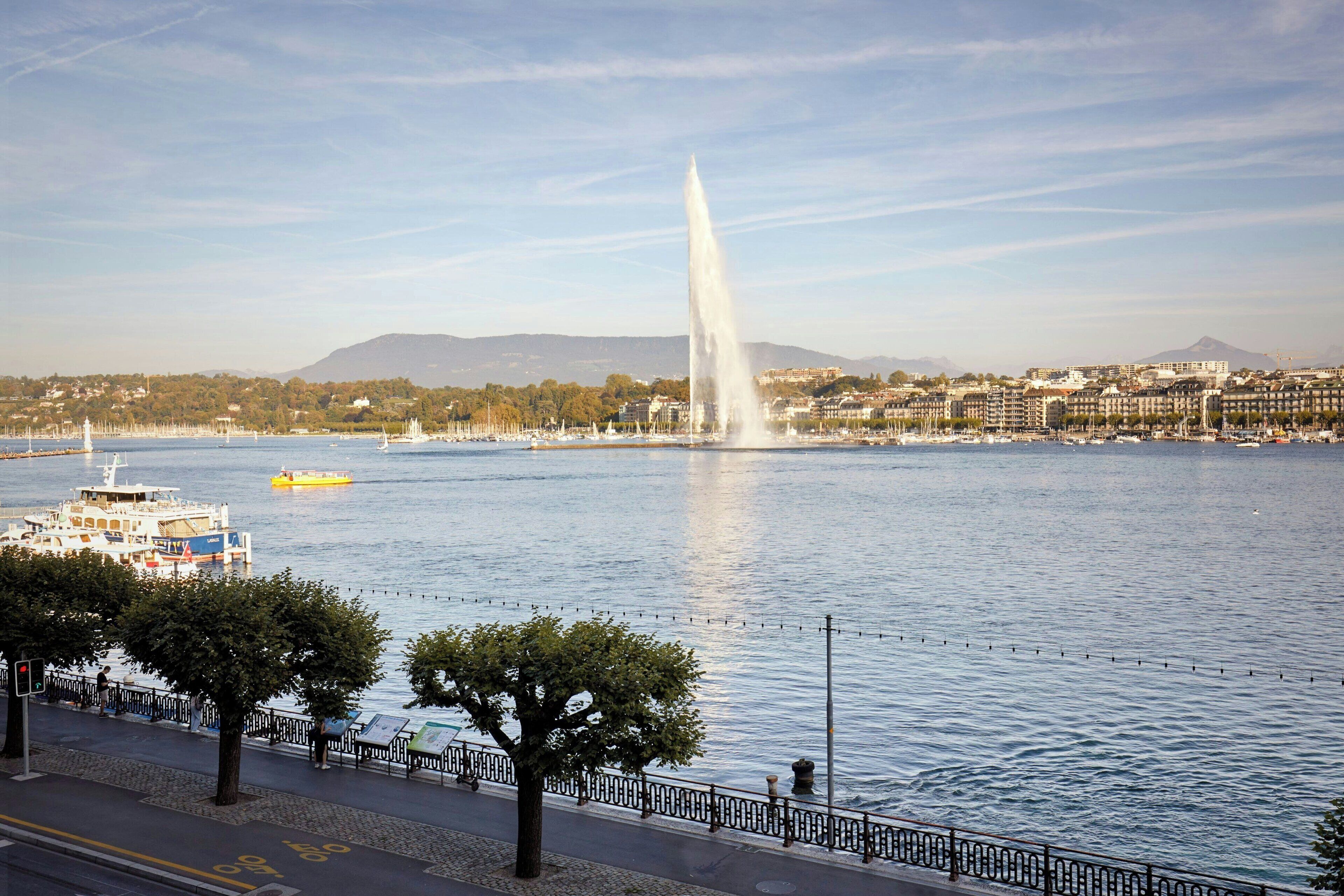 A waterfront cityscape with a prominent fountain, a boat docked on the river, and a pedestrian walkway along the water's edge.