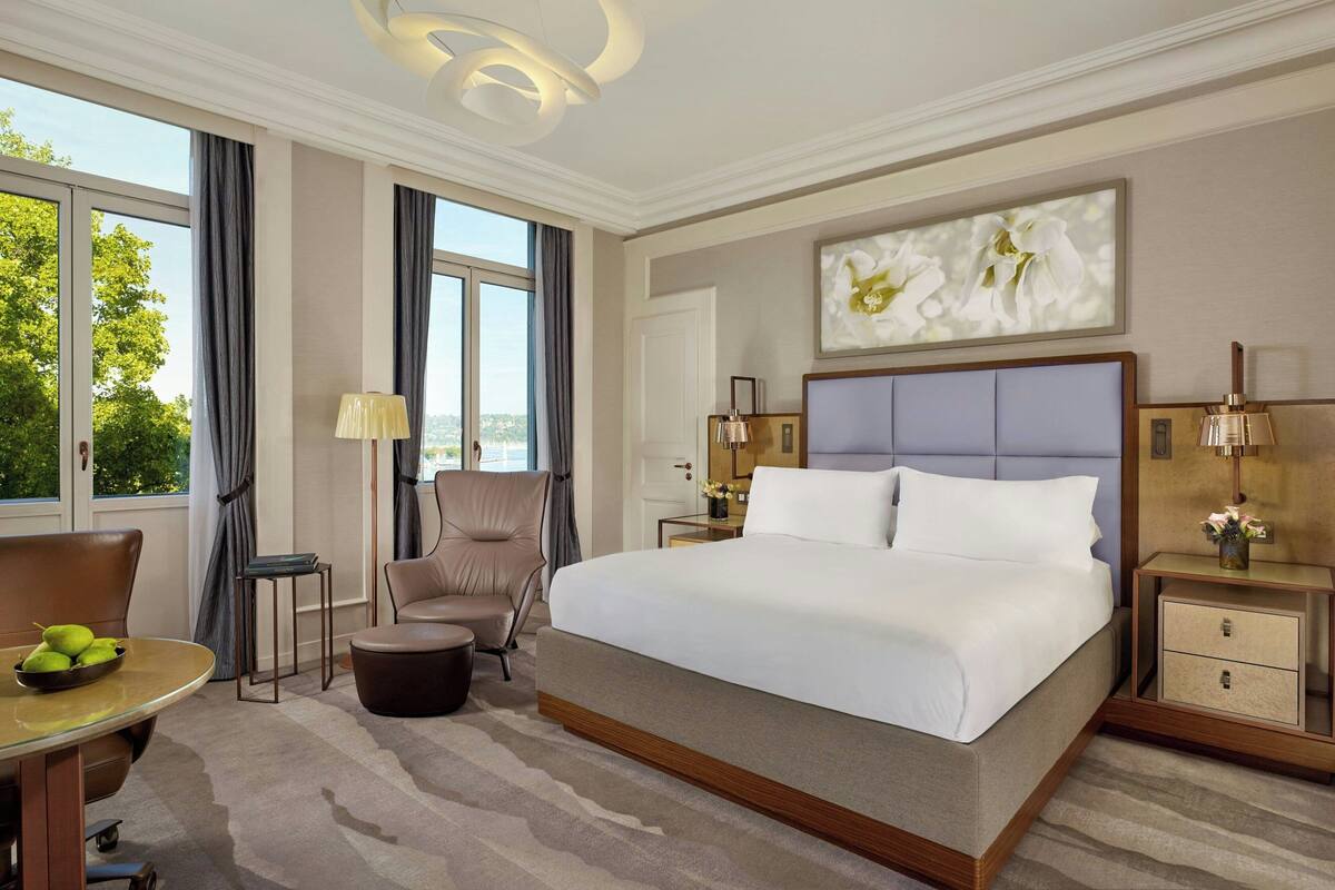 A modern hotel room with a large bed, a sitting area with a table and chairs, a small side table with a lamp, and a view of the outdoors.