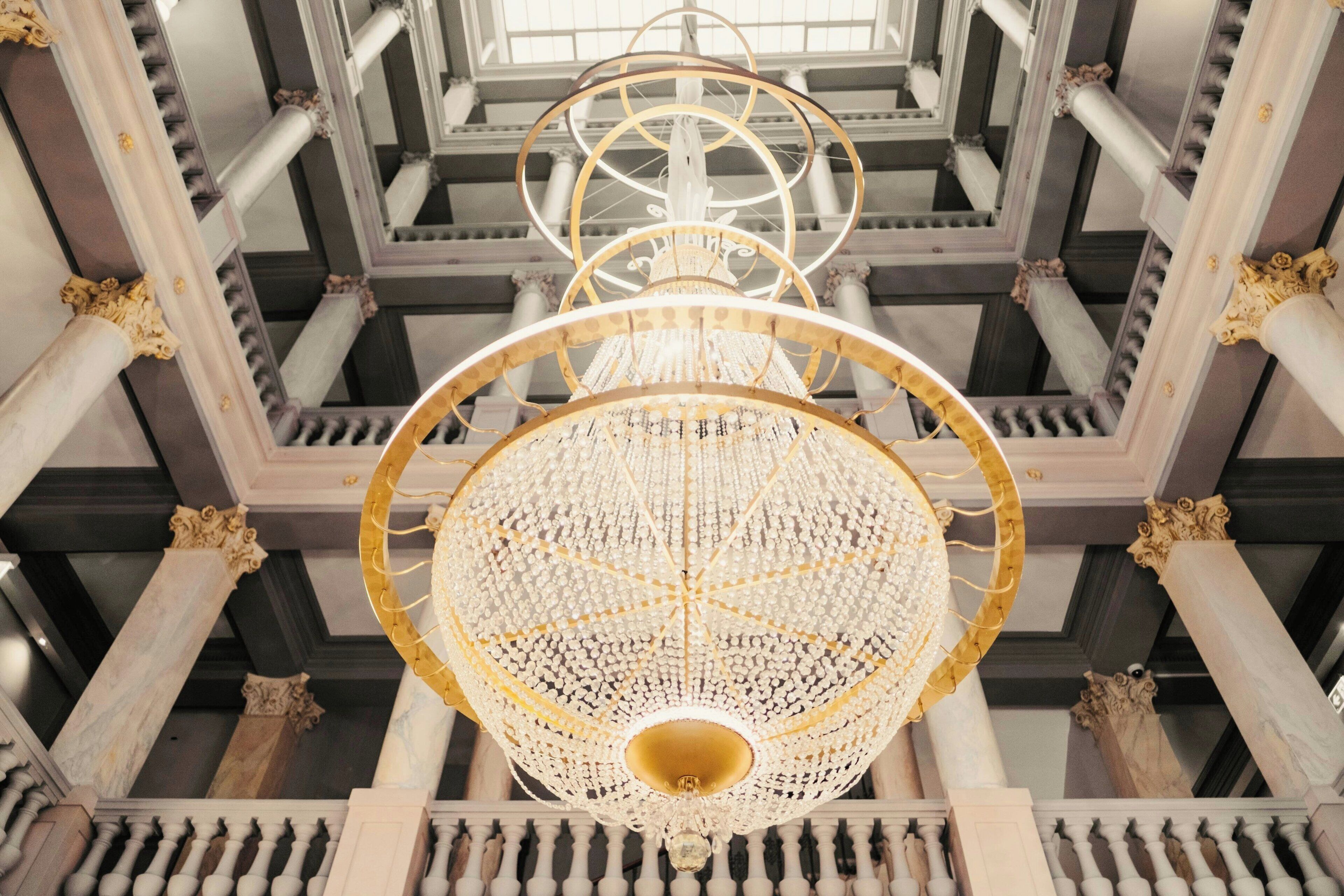 A grand chandelier with multiple tiers and intricate details, set in a spacious hall with classical architecture and marble columns.