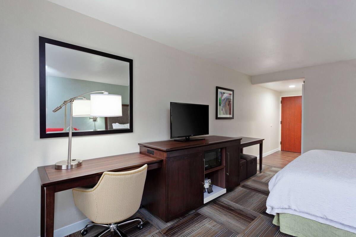 A hotel room with a bed, a desk with a chair, a television, a mirror, and a door leading to another room.