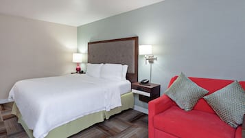 A hotel room with a bed, bedside table, lamp, and a red sofa with patterned cushions.