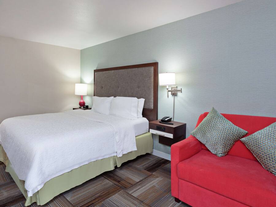 A hotel room with a bed, bedside table, lamp, and a red sofa with patterned cushions.