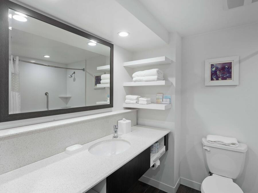 A modern bathroom with a large mirror, white sink, and toilet. There are shelves with towels and toiletries.