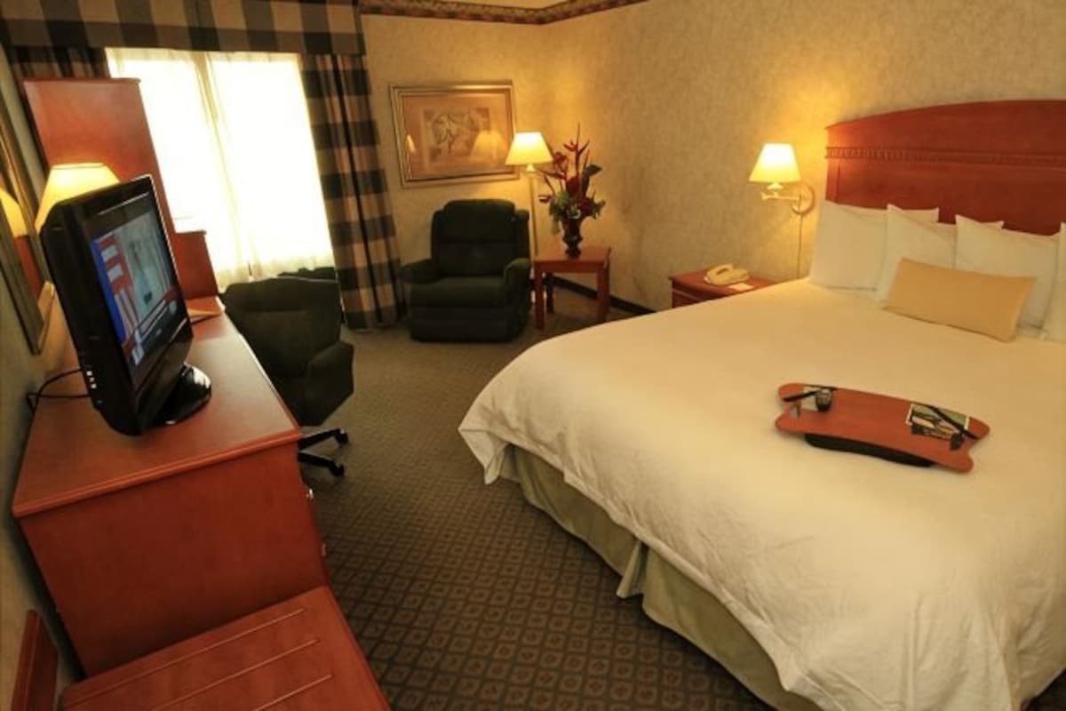 A hotel room with a bed, a desk with a TV, a chair, a nightstand with a lamp, and a painting on the wall.
