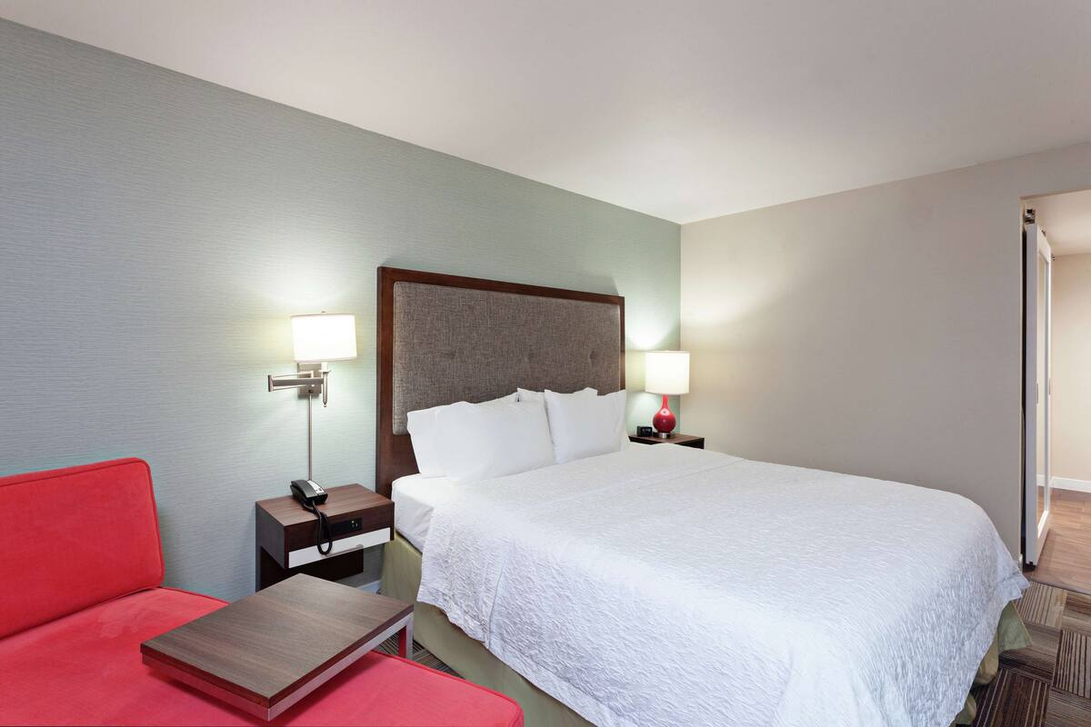A hotel room with a large bed, a red armchair, a wooden nightstand with a lamp, and a small table.