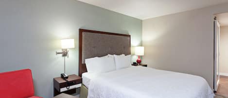 A hotel room with a large bed, a red armchair, a wooden nightstand with a lamp, and a small table.