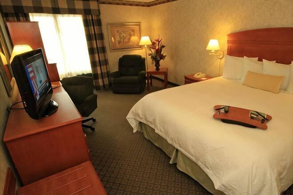 A hotel room with a bed, a desk with a TV, a chair, a nightstand with a lamp, and a painting on the wall.
