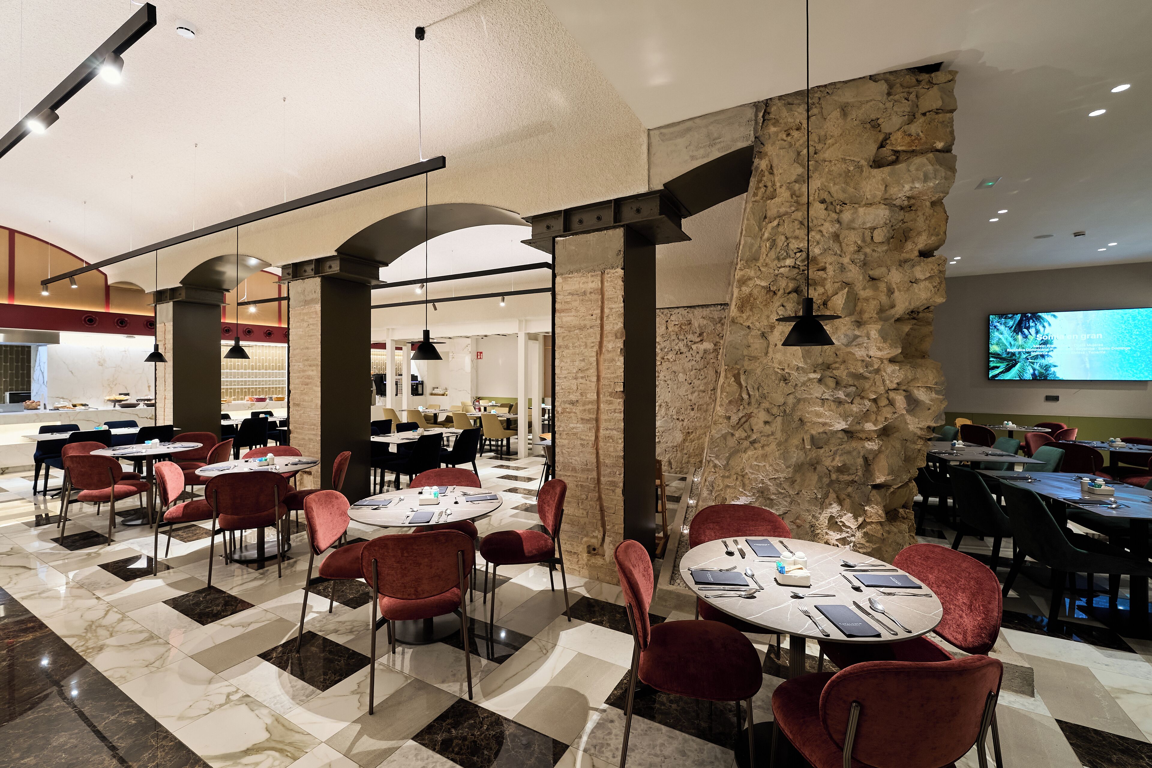 A spacious dining area with red chairs and round tables, featuring a stone wall and modern pendant lighting.