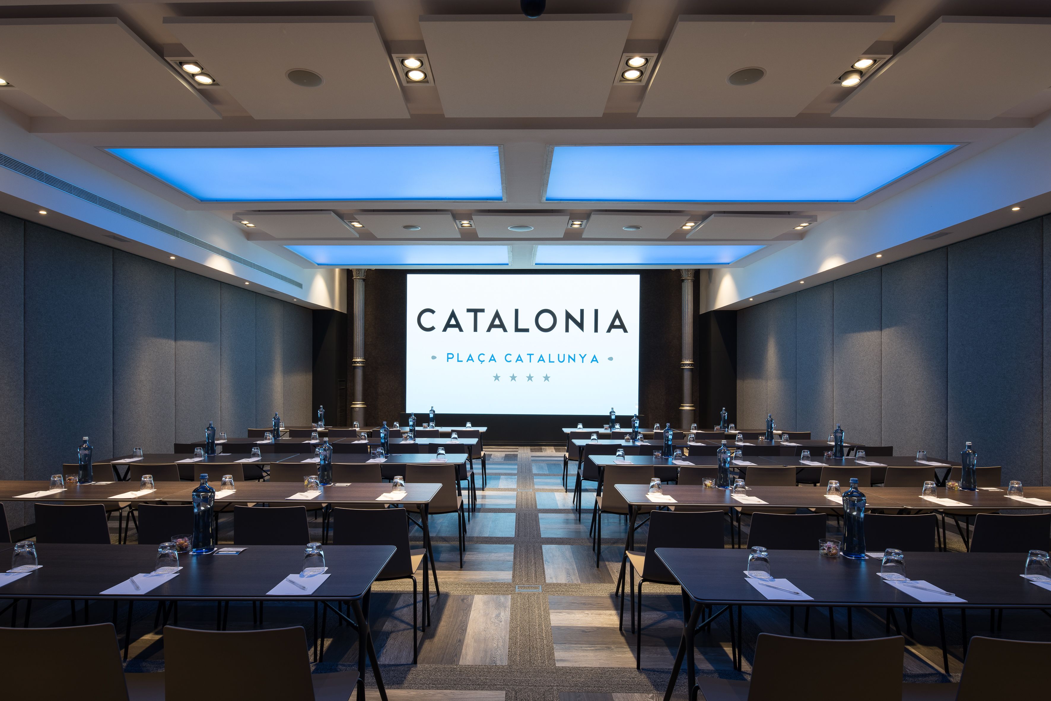 Une salle de conférence dotée de plusieurs tables et chaises, d’un grand écran affichant la mention « CATALONIE, PLACE DE CATALOGNE », et d’un éclairage encastré.