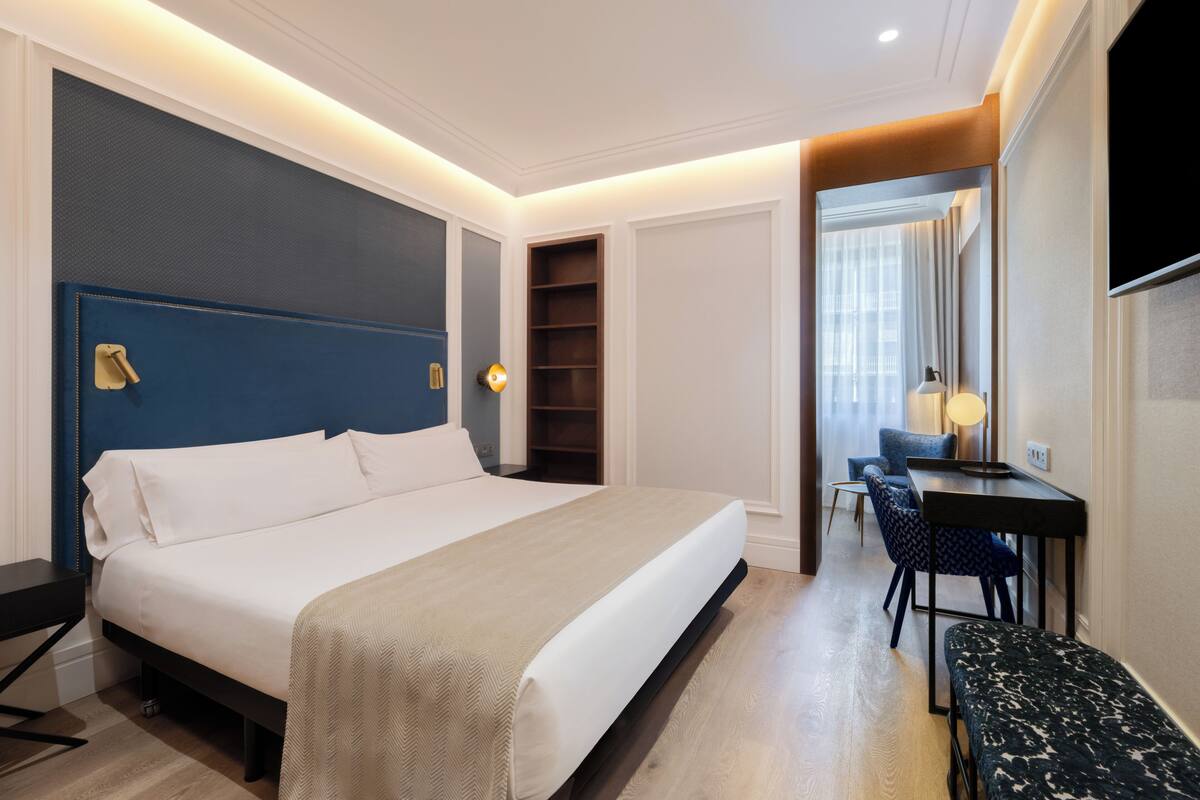 Quarto de hotel com uma cama grande, mesas de cabeceira, escrivaninha com cadeira, um pequeno espaço de estar e uma televisão.