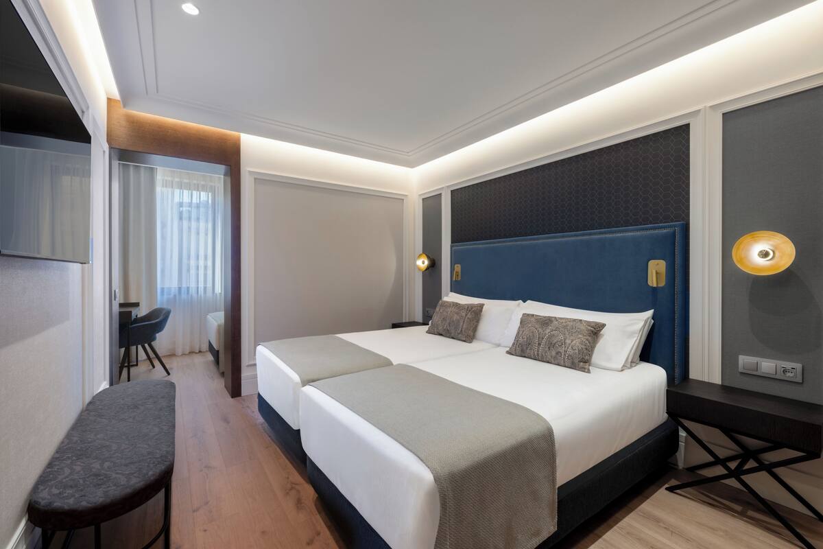 Quarto de hotel com duas camas, uma escrivaninha, um banco e uma televisão.
