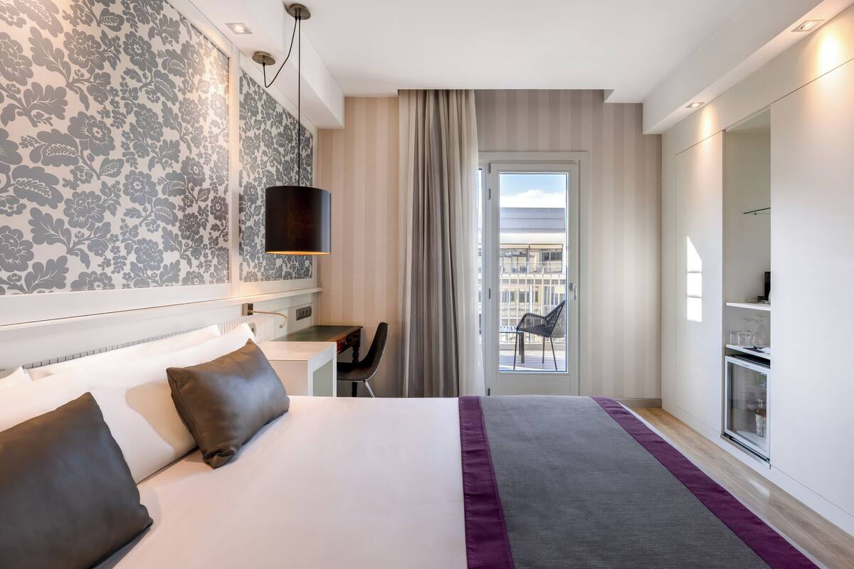 Quarto de hotel com cama, escrivaninha, cadeira e vista para a cidade pela janela.