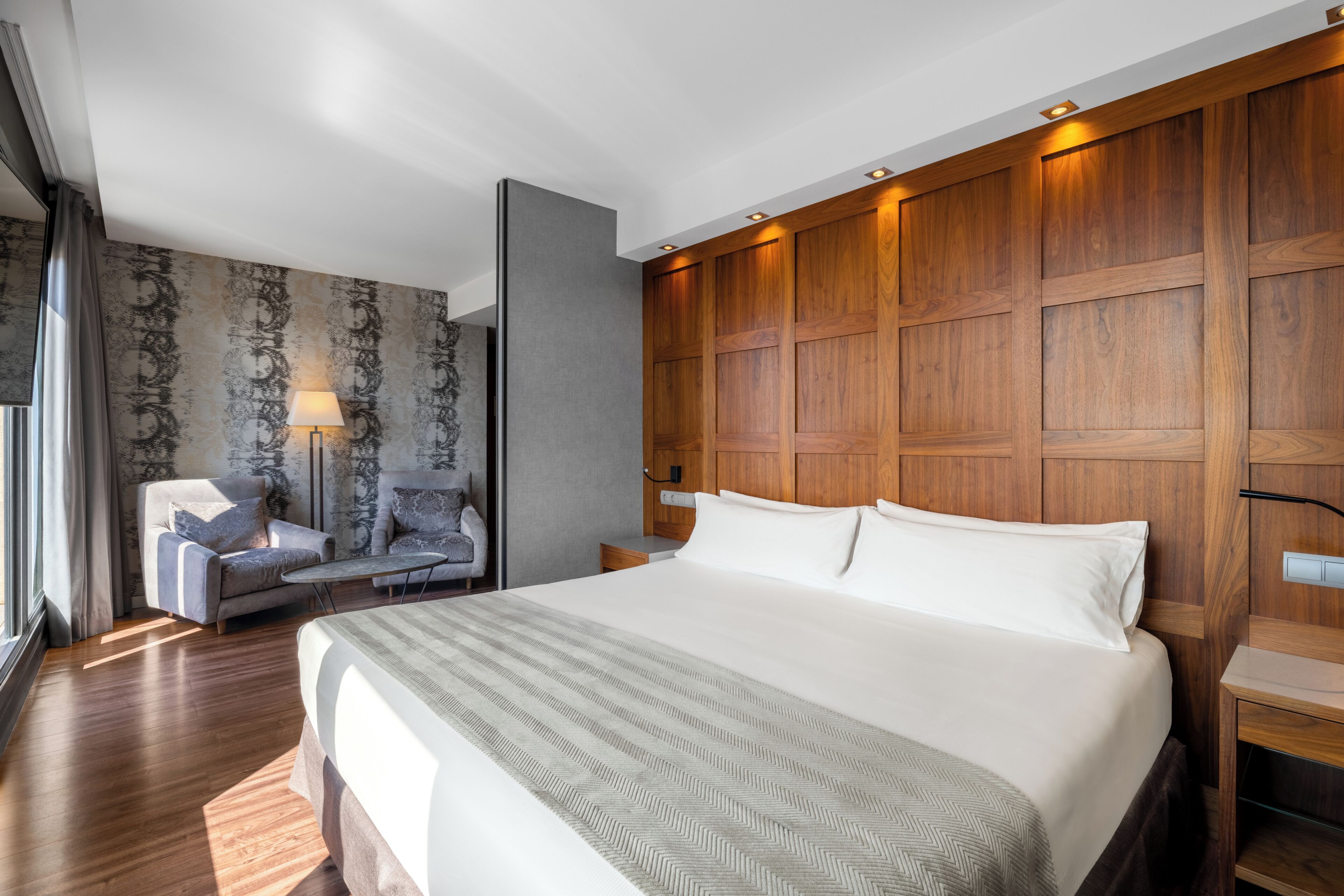 A modern hotel room with a large bed, a seating area with a sofa and armchair, a wooden headboard, and a patterned wallpaper wall.