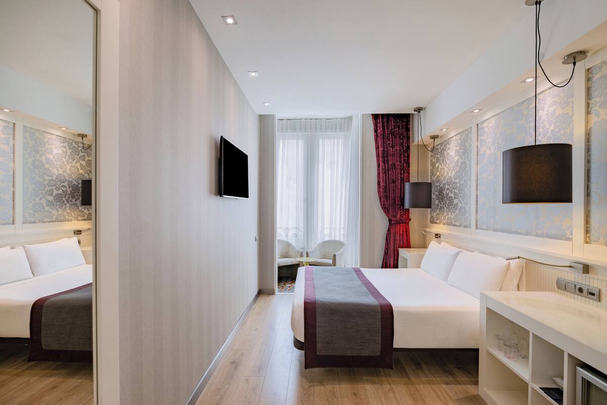 Quarto de hotel com duas camas, televisão, espelho, escrivaninha e janela com cortinas.