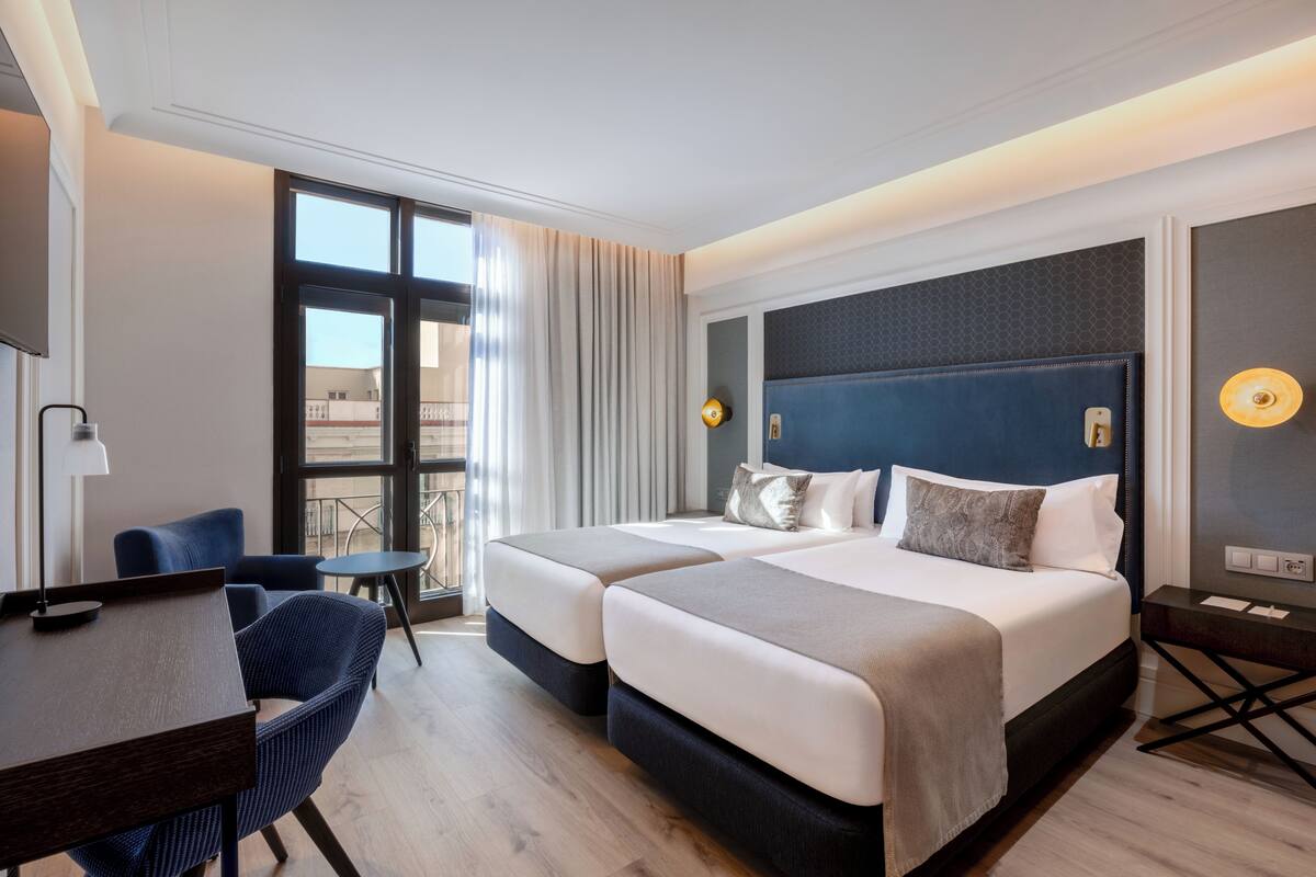 Quarto de hotel com duas camas, uma escrivaninha, uma cadeira e vista para o exterior.
