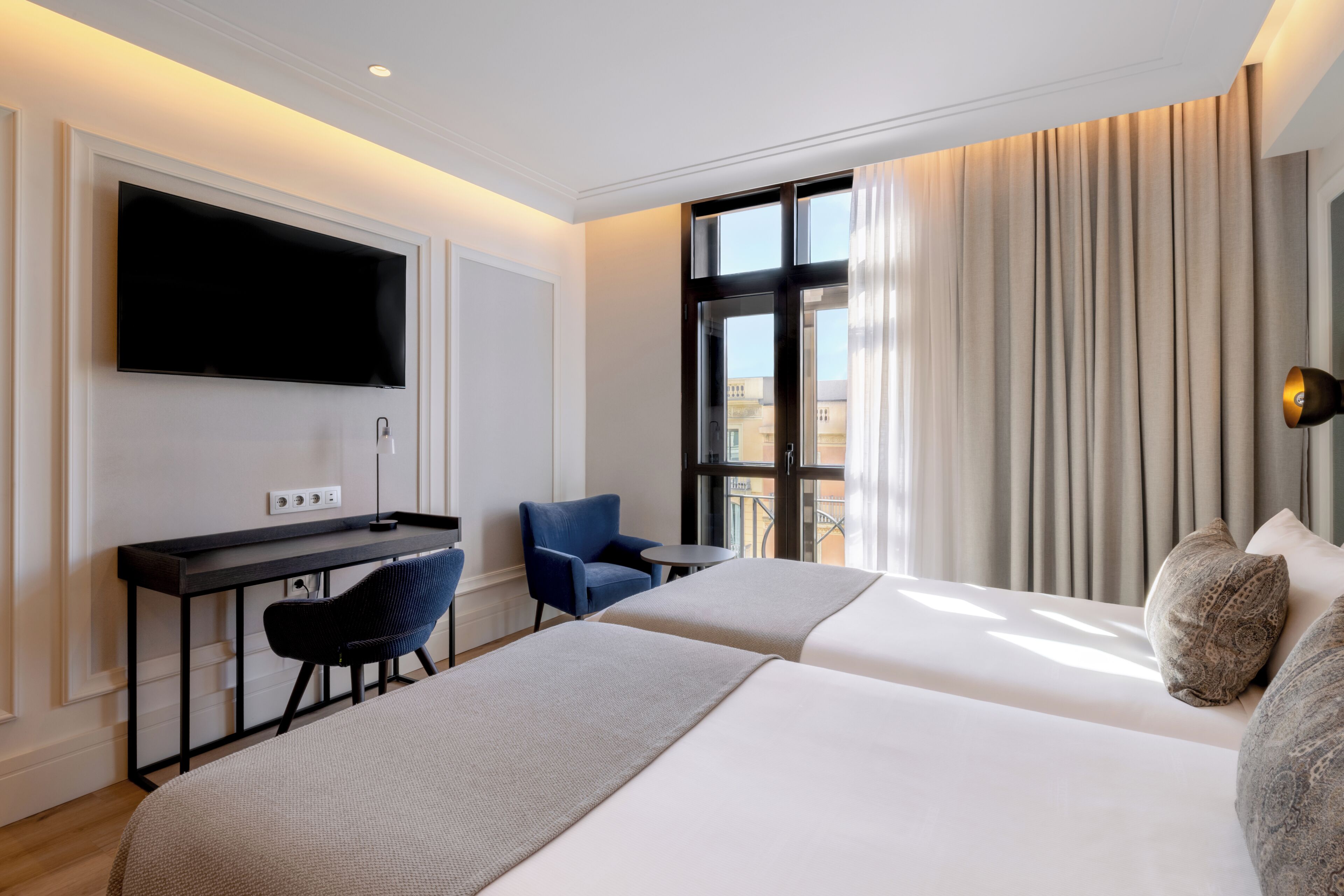 Habitación de hotel con dos camas, un escritorio con silla, televisor y ventana con cortinas.
