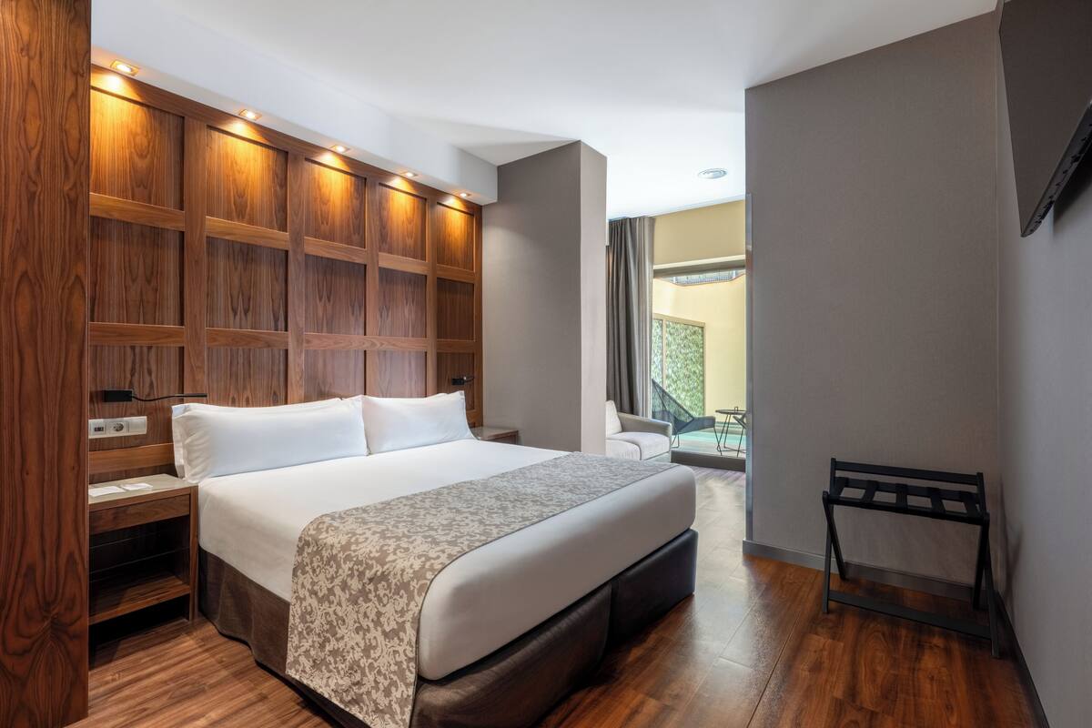 Quarto de hotel com uma cama grande, painéis de madeira, um pequeno espaço de estar e vista para o exterior.