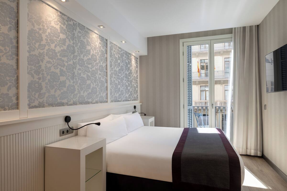 Quarto de hotel com uma cama grande, uma mesa de cabeceira, televisão na parede e vista para uma varanda com edifícios.