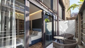 Moderni hotellihuone, jossa on lasiovi, sänky, pieni pöytä ja punotut tuolit.