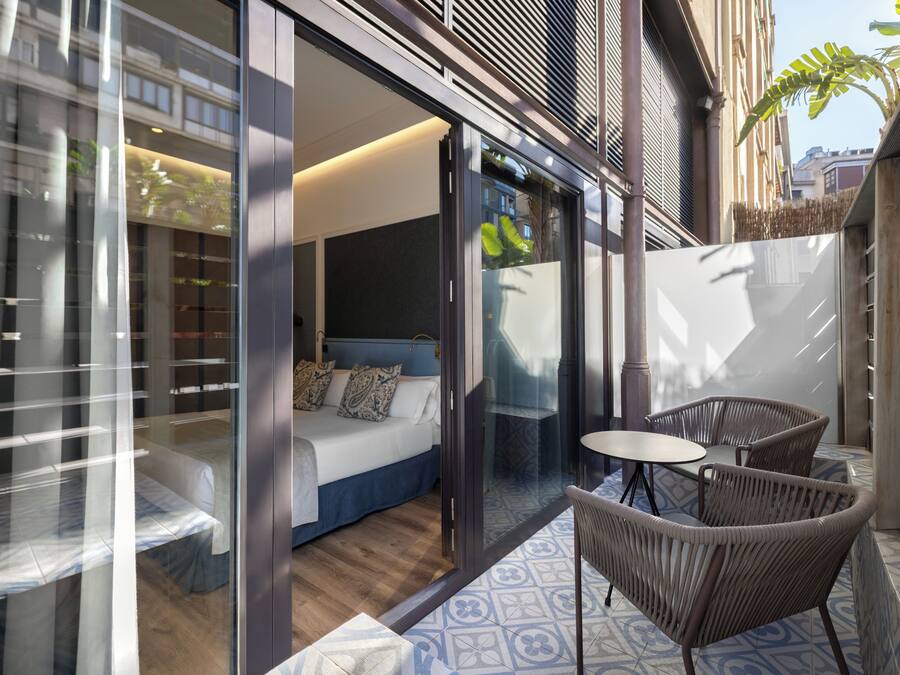 Una habitación de hotel moderna con puerta de vidrio, una cama, una mesita pequeña y sillas de mimbre.
