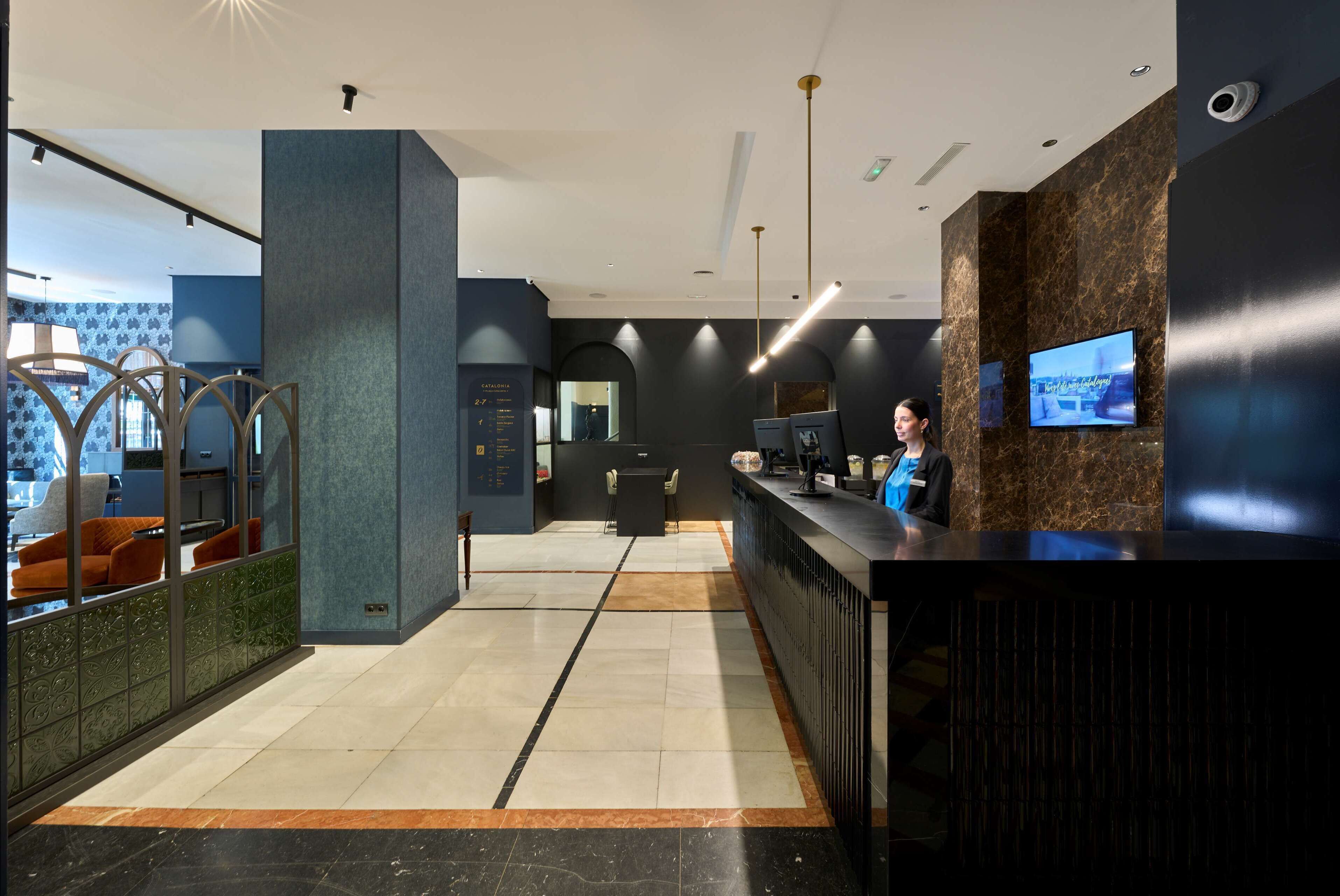 A modern reception area with a marble floor, a reception desk, a TV screen, and a seating area with orange chairs.