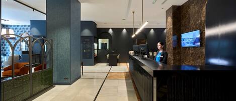 A modern reception area with a marble floor, a reception desk, a TV screen, and a seating area with orange chairs.