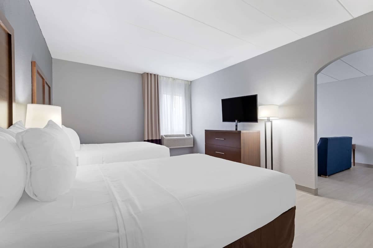 A hotel room with two beds, a flat-screen TV, a desk with a lamp, and a blue seating area.