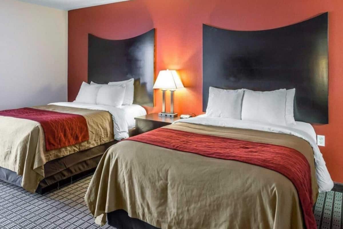 A hotel room with two beds, a nightstand with a lamp, and a wall with a large black panel.