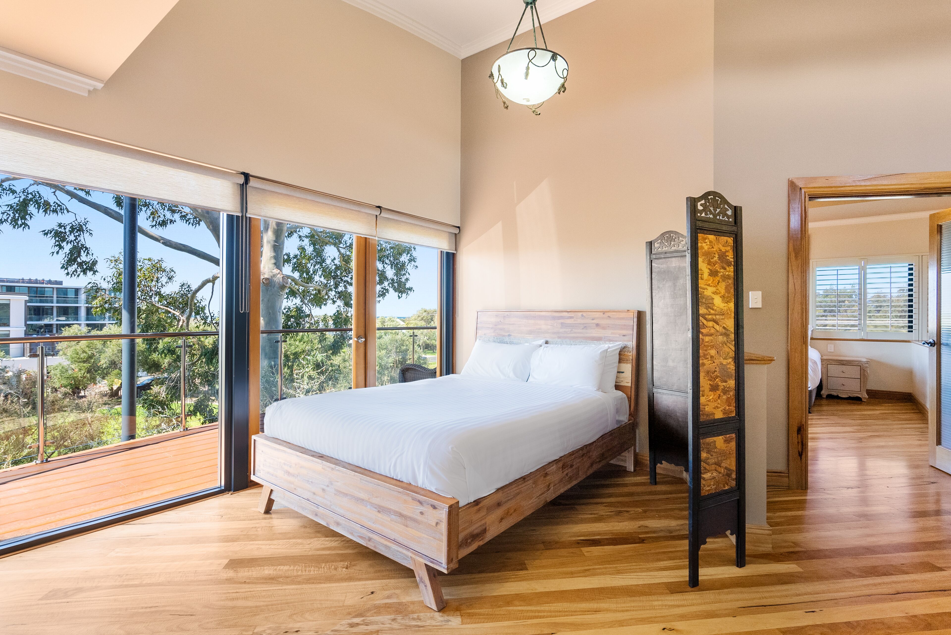 A bedroom with a large bed, a wooden floor, a view of greenery, and a ceiling light.