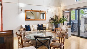 A modern living room with a glass-top dining table surrounded by wicker chairs, a sofa with cushions, a large mirror, and a potted plant. - Bay Village Resort Dunsborough (Dunsborough)