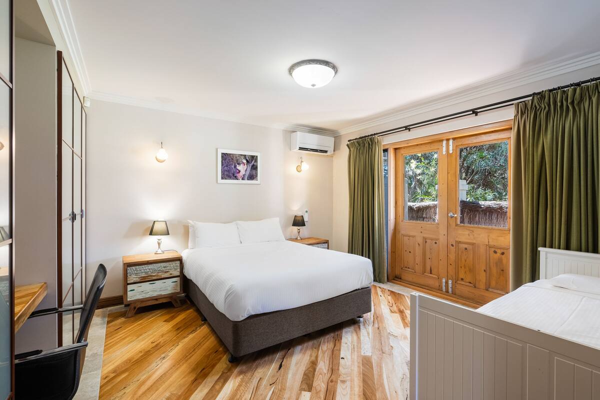 A bedroom with a wooden floor, a bed, a nightstand, a lamp, a window with curtains, and a wall-mounted air conditioner.