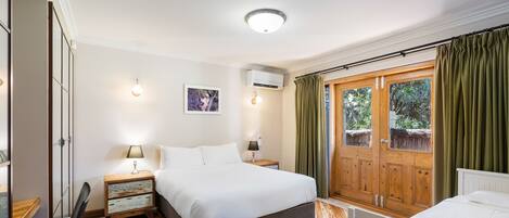 A bedroom with a wooden floor, a bed, a nightstand, a lamp, a window with curtains, and a wall-mounted air conditioner.