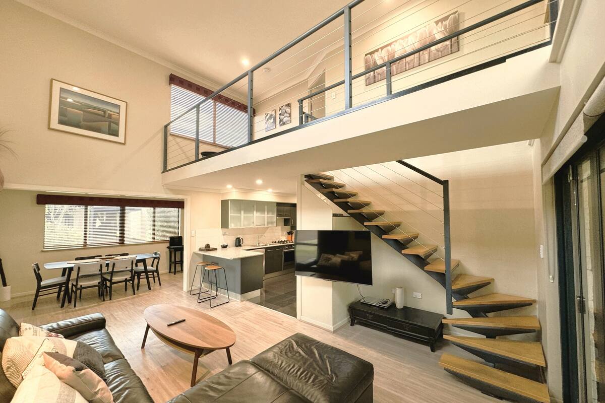 A spacious living area with a high ceiling, a staircase, a kitchen, and a dining area.