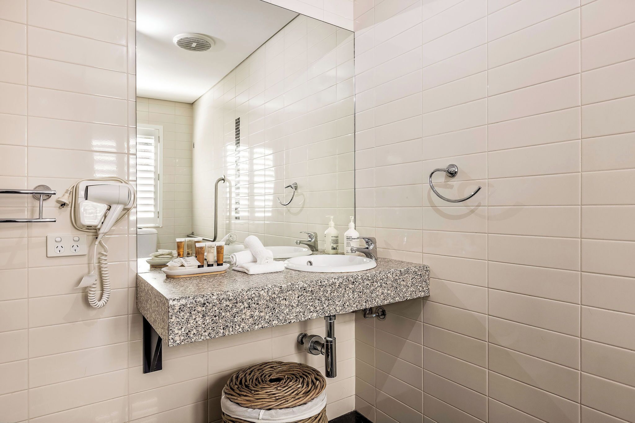 A bathroom with white tiled walls, a granite countertop, a sink, a mirror, a hairdryer, a towel, and a waste basket.