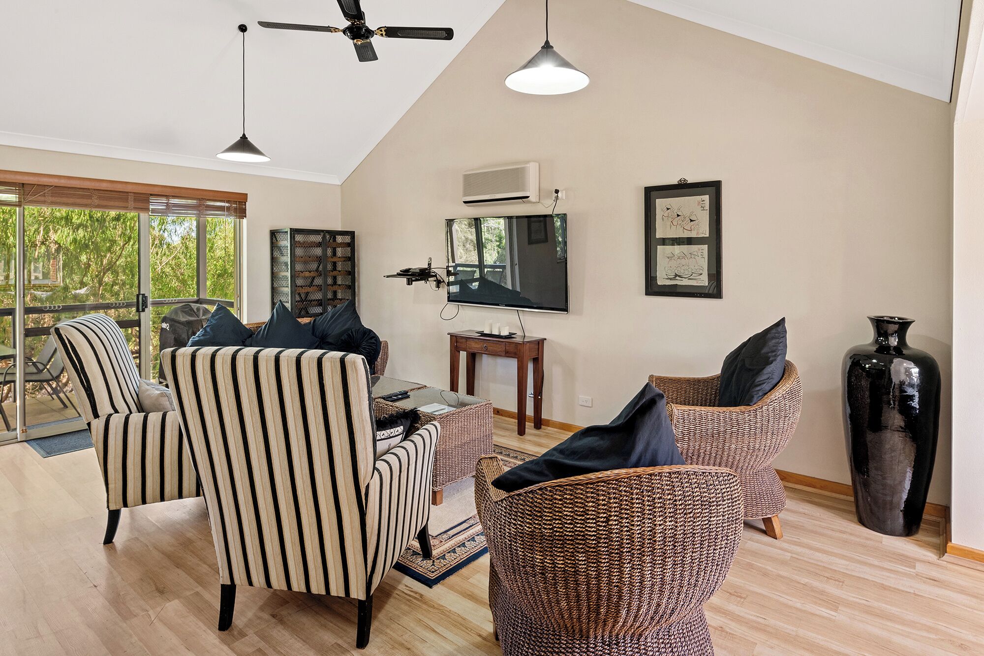 Un salon spacieux avec une grande télévision, des fauteuils à rayures, des canapés en osier et un ventilateur de plafond.