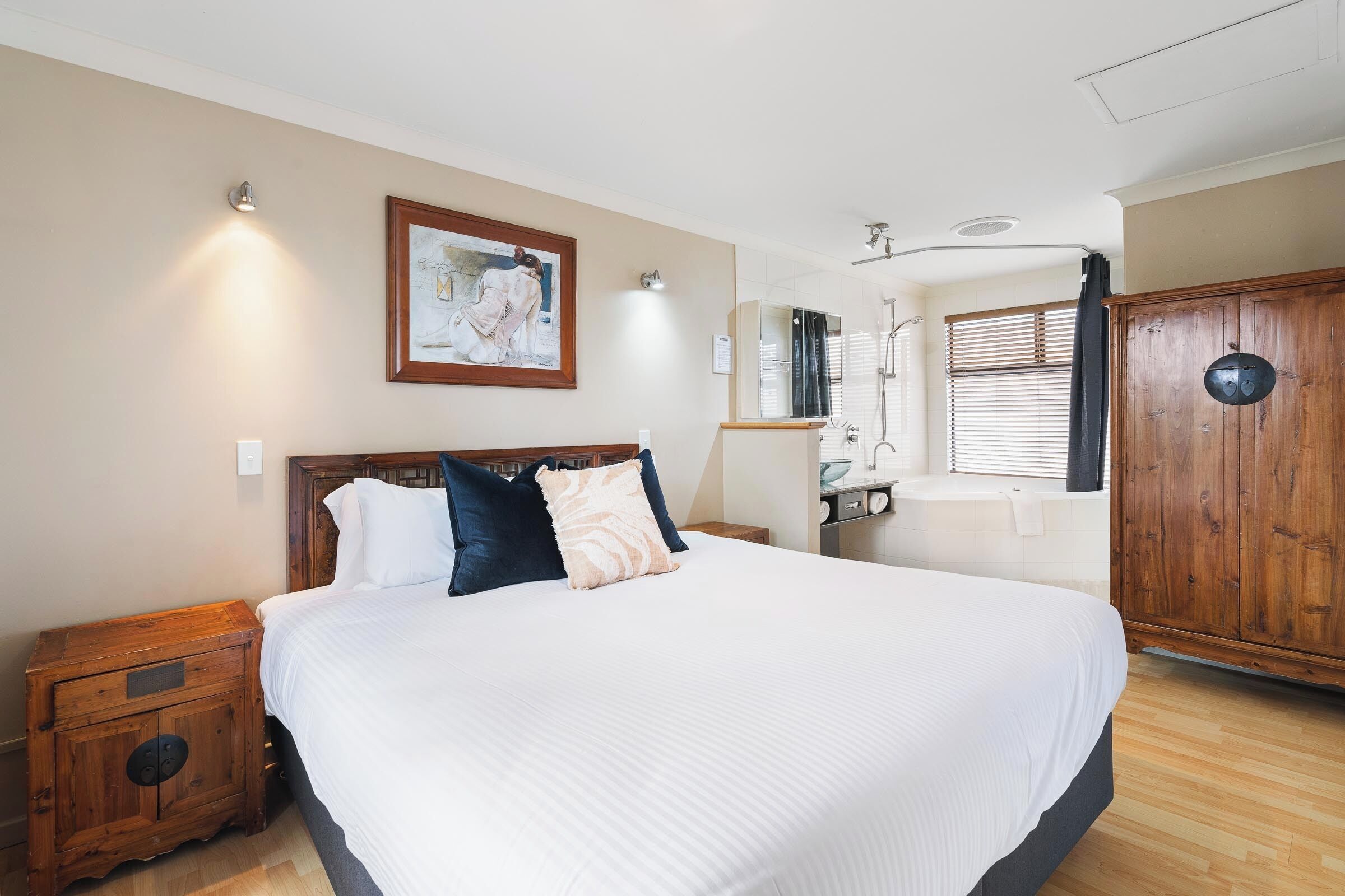 A large bed with white bedding, a wooden nightstand, a wooden wardrobe, and a bathroom with a bathtub and shower.