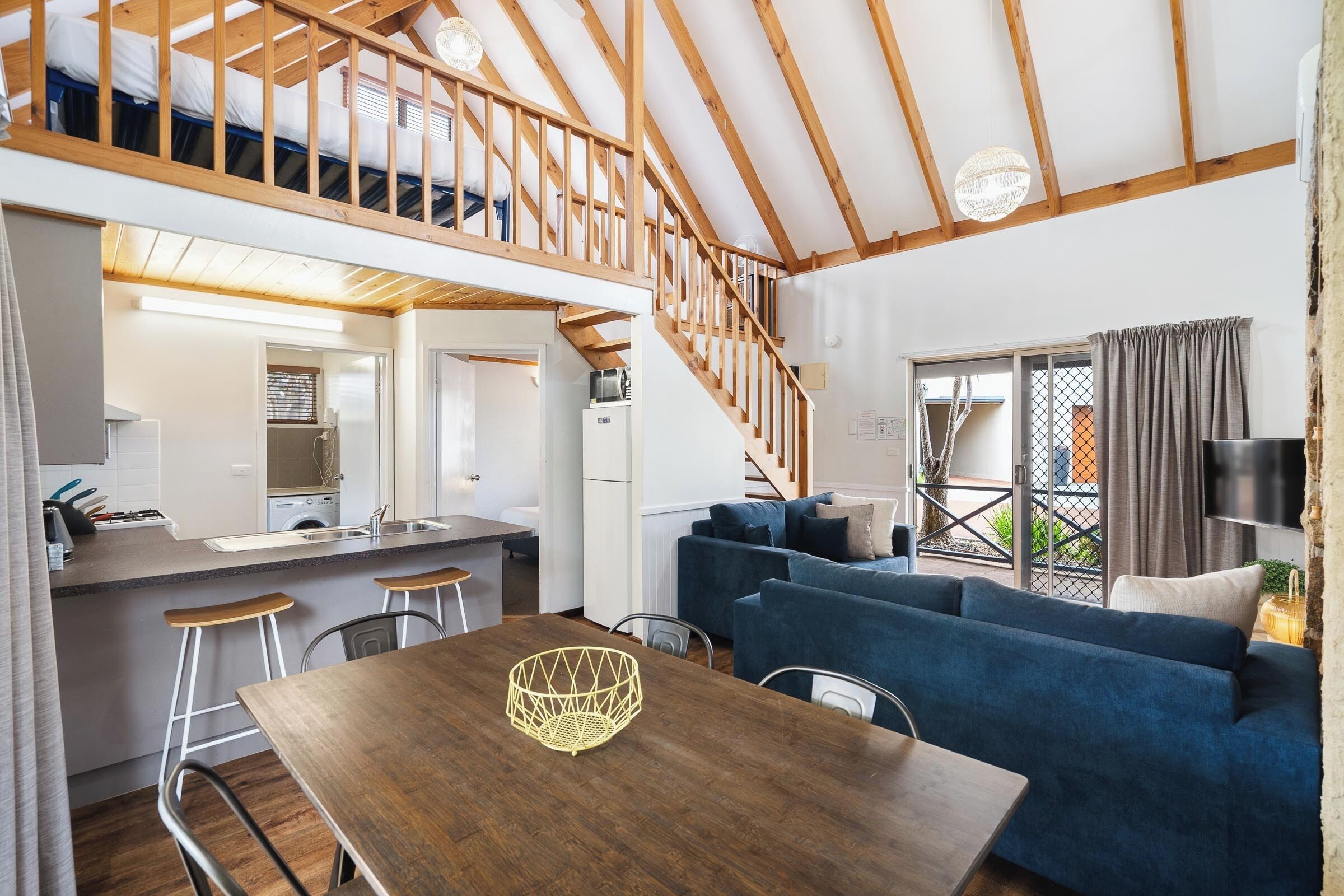 A modern loft-style apartment with a wooden staircase, a kitchen with a sink and bar stools, a dining area with a wooden table, and a living area with a blue sofa.