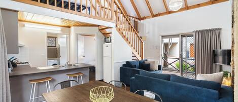 A modern loft-style apartment with a wooden staircase, a kitchen with a sink and bar stools, a dining area with a wooden table, and a living area with a blue sofa.