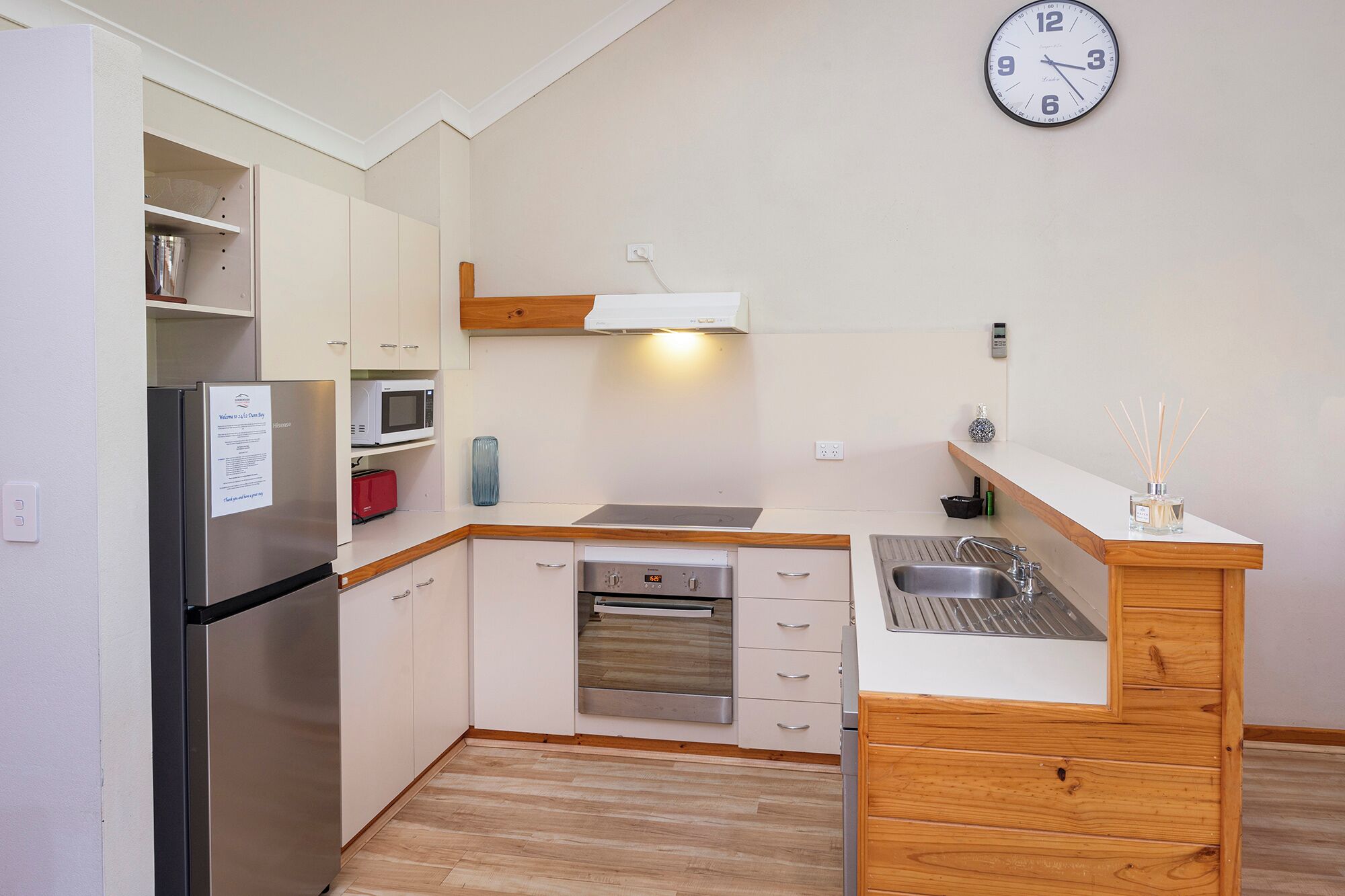 Cocina compacta con refrigerador, microondas, horno y muebles de madera. Un reloj está colgado en la pared.