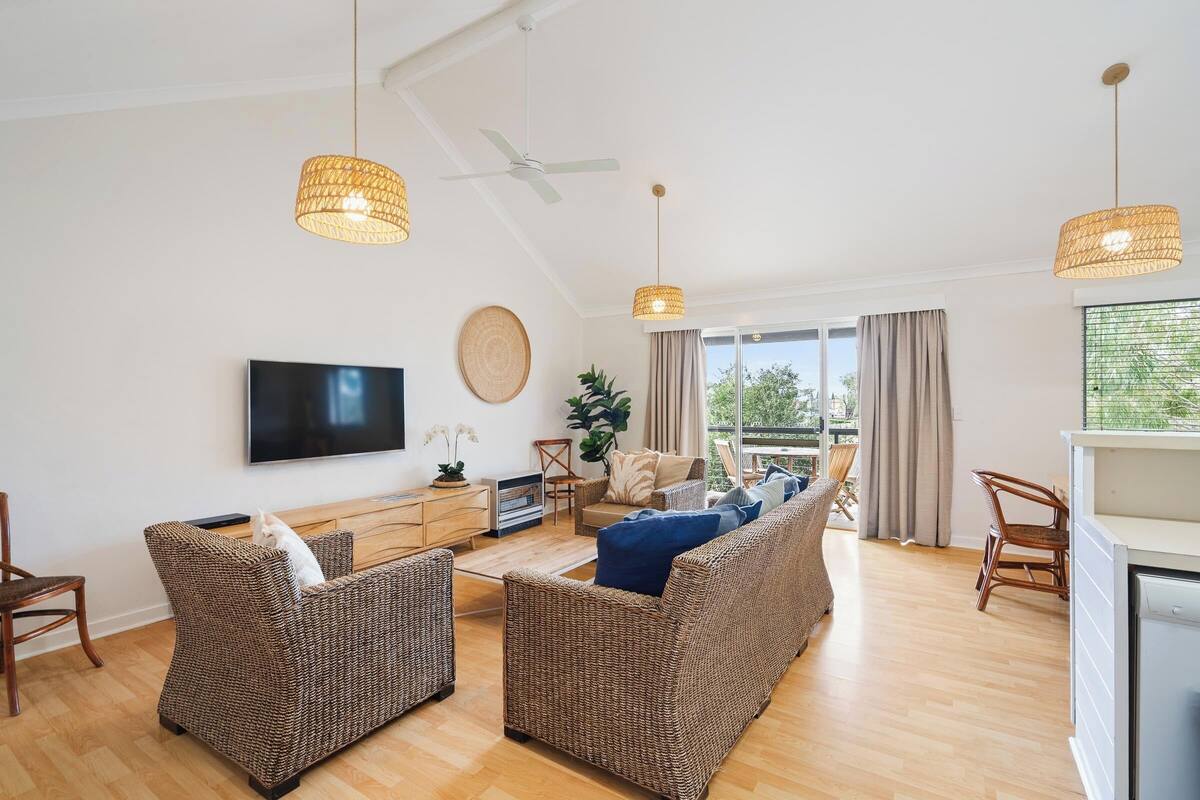 A modern living room with wicker furniture, a flat-screen TV, and large windows offering a view of the outdoors.