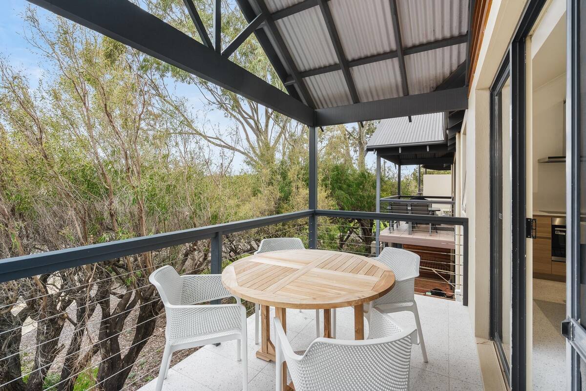 A covered outdoor patio with a round wooden table and white wicker chairs, surrounded by a metal railing.