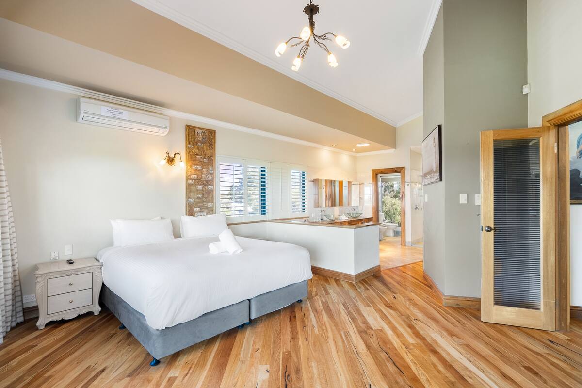 A modern bedroom with a large bed, wooden flooring, a built-in desk, and a bathroom visible through an open door.
