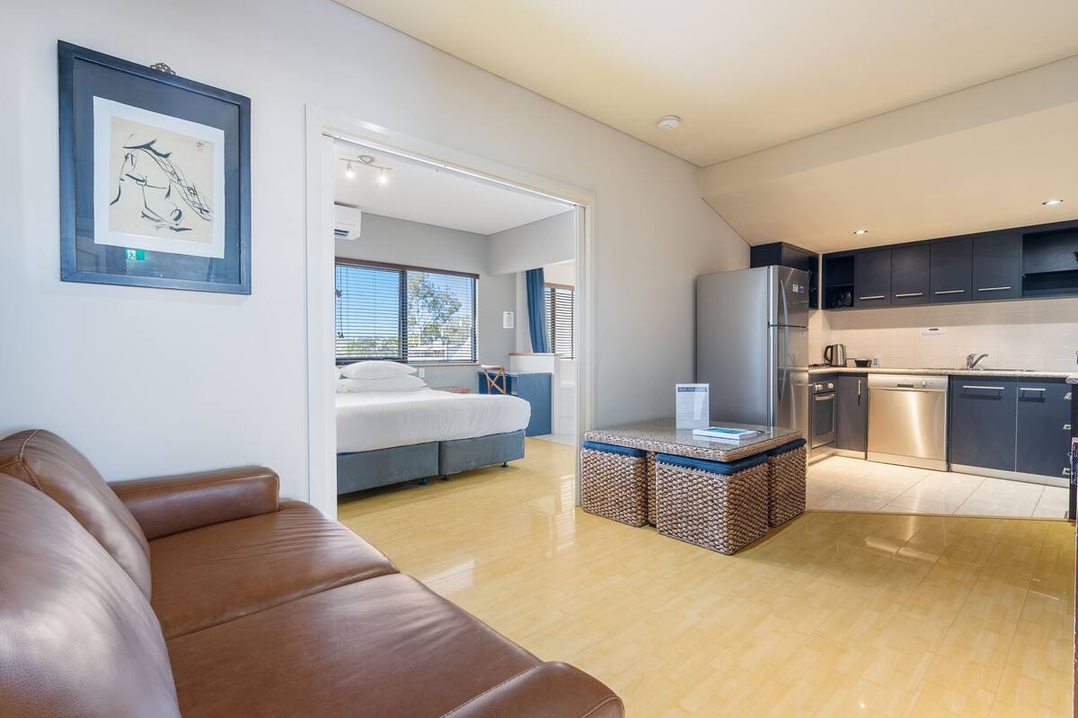 A modern living room with a leather sofa, a bed with a blue headboard, and a kitchen with dark cabinets and stainless steel appliances.