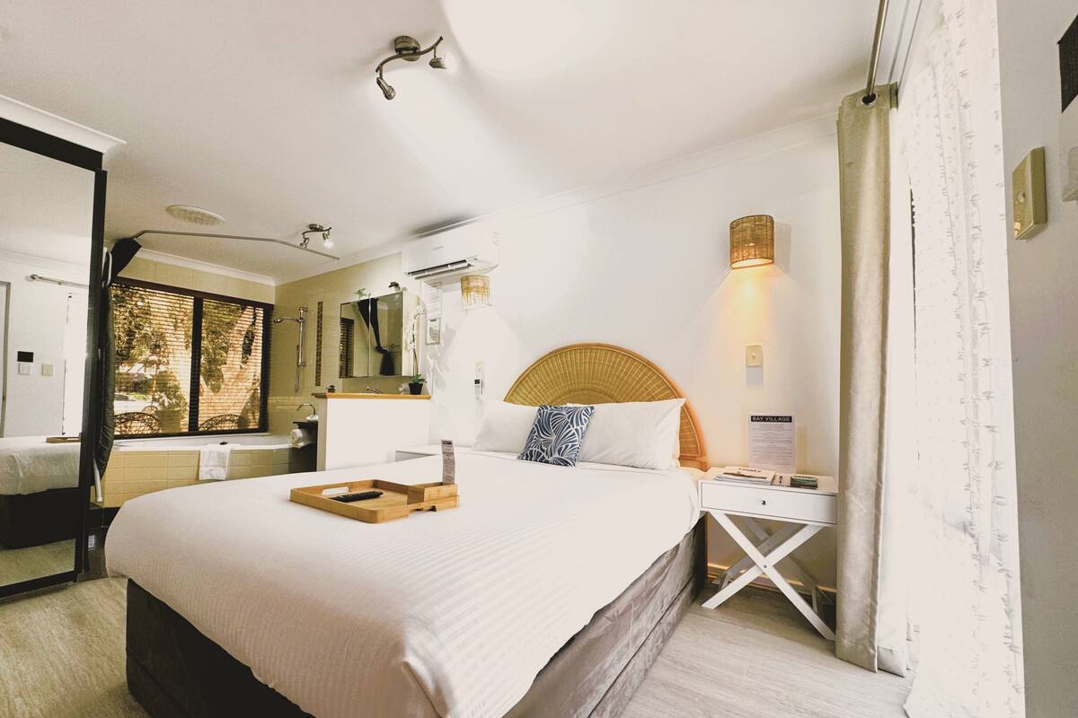 A hotel room with a large bed, a bedside table with a lamp, a window with blinds, a bathroom visible through the door, and a sign on the wall.