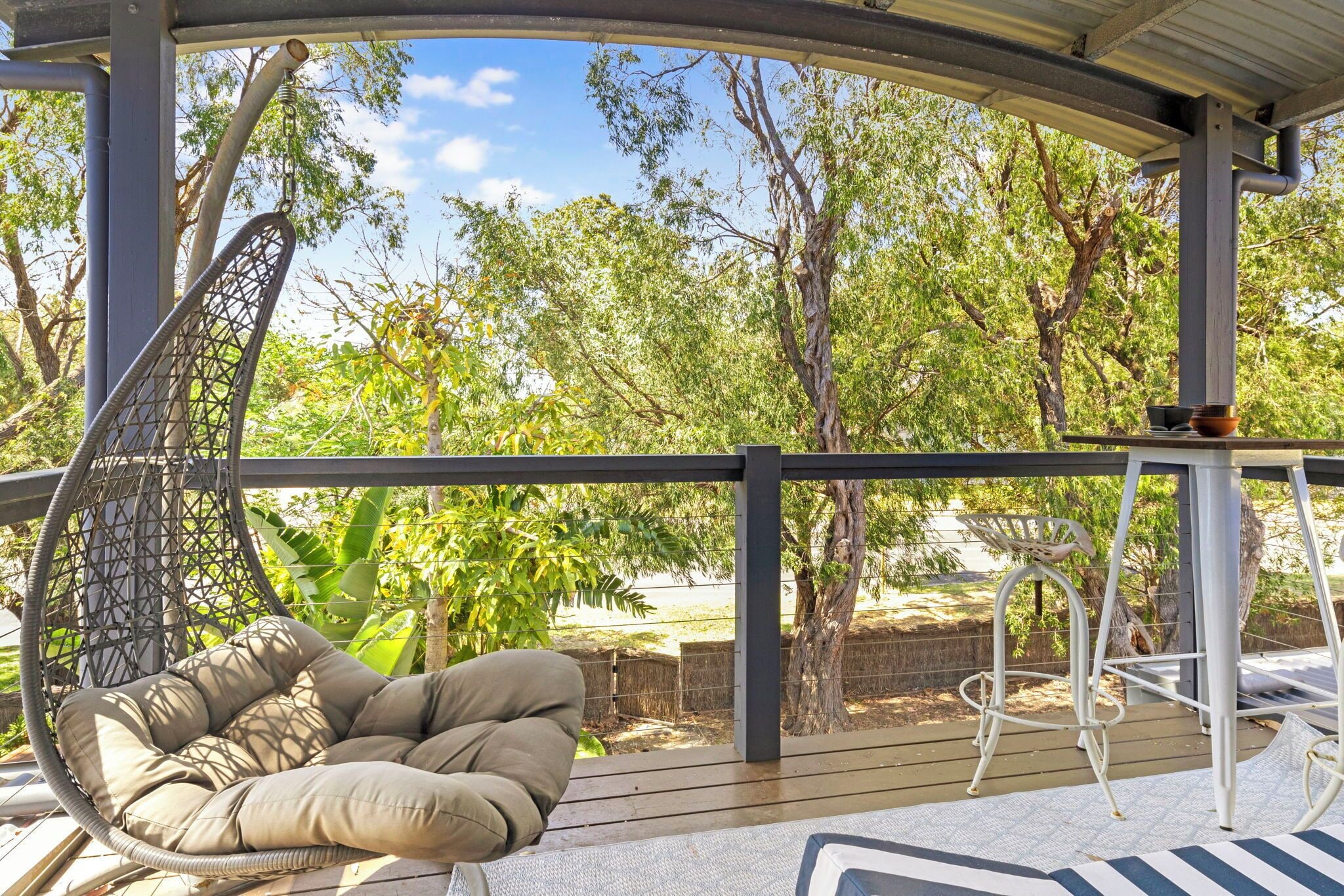 A covered outdoor area with a hanging chair, a table with a bowl, and a view of trees and a fence.
