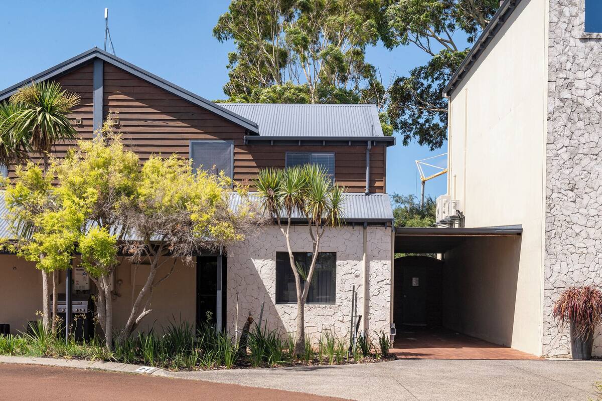 A two-story house with a garage, a stone facade, and a variety of vegetation including trees and shrubs.