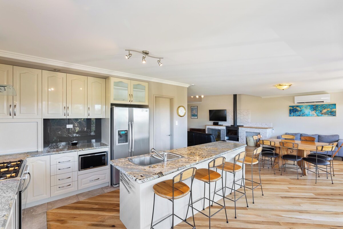 Una cocina moderna con una isla central, taburetes de bar y una zona de comedor con mesa y sillas de madera.